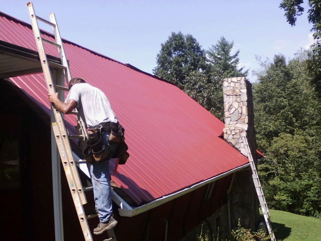 Roofer on a ladder beside a red metal roof and stone chimney — Soffit & Fascia Repair by Roofing Brothers in Broadview